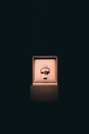 A single ring with a circular gemstone is elegantly displayed in an illuminated jewelry box set against a dark background. Soft lighting enhances the ring's sparkle and creates a luxurious atmosphere.