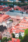 An aerial view of a picturesque European town features tightly packed historical buildings with red-tiled roofs at the forefront. The square hosts people walking and socializing, and a river bordered by greenery flows in the background. Across the river, brightly colored buildings complete the charming scenery, along with a bridge connecting the two sides.