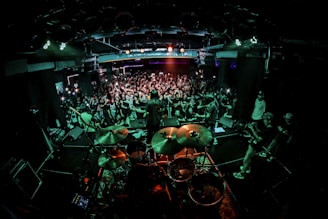 A live concert scene with a band performing on stage. The drummer is prominent in the foreground, and the audience is densely packed, eagerly watching and capturing the moment with cameras and phones. Bright stage lights illuminate the venue, creating a dynamic and energetic atmosphere.