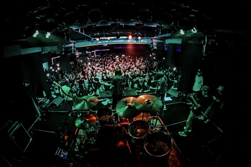 A live concert scene with a band performing on stage. The drummer is prominent in the foreground, and the audience is densely packed, eagerly watching and capturing the moment with cameras and phones. Bright stage lights illuminate the venue, creating a dynamic and energetic atmosphere.