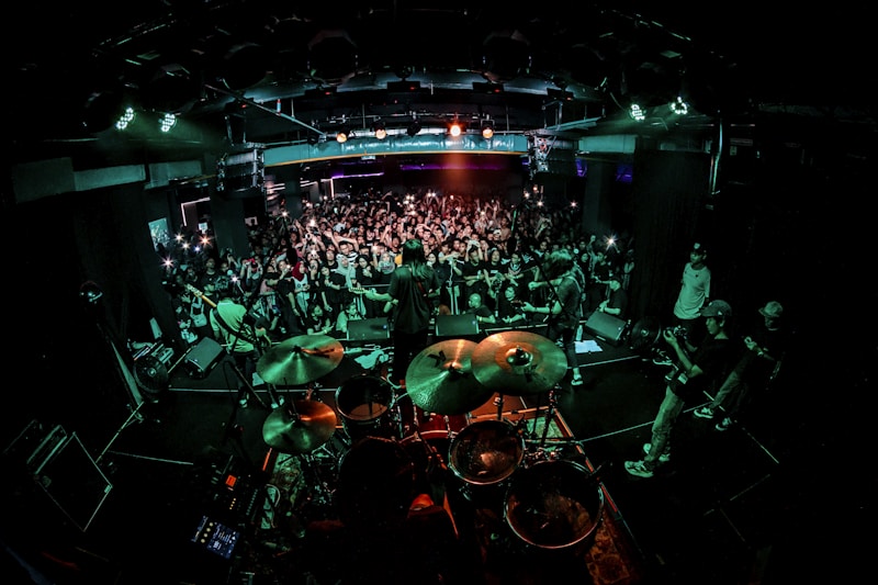 A live concert scene with a band performing on stage. The drummer is prominent in the foreground, and the audience is densely packed, eagerly watching and capturing the moment with cameras and phones. Bright stage lights illuminate the venue, creating a dynamic and energetic atmosphere.