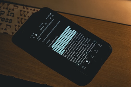A smartphone is placed on a wooden surface displaying a text in a dark mode setting, suggesting reading or studying. The background features a partial view of a patterned surface, possibly paper or a book cover.