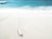 A white tube of sunscreen lies on a sandy beach near the shore, with gently crashing waves and turquoise water in the background.
