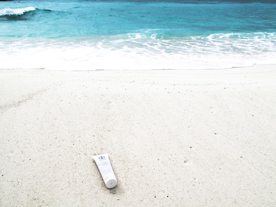 A white tube of sunscreen lies on a sandy beach near the shore, with gently crashing waves and turquoise water in the background.