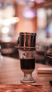 A Vietnamese coffee drip filter resting on top of a glass containing a dark and layered coffee beverage. The background is softly blurred with warm lighting, creating a cozy ambiance.