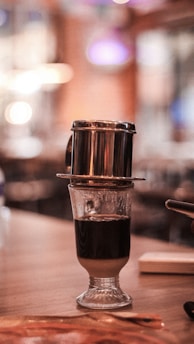 A Vietnamese coffee drip filter resting on top of a glass containing a dark and layered coffee beverage. The background is softly blurred with warm lighting, creating a cozy ambiance.