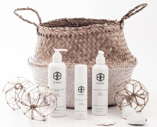A woven basket with handles holds three cylindrical skincare product bottles labeled ONNE, which include a pump bottle, a spray bottle, and a smaller cream bottle. Around the basket are two decorative spherical ornaments made of glass and twine, and some smooth stones.