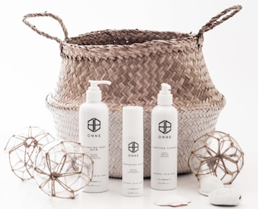 A woven basket with handles holds three cylindrical skincare product bottles labeled ONNE, which include a pump bottle, a spray bottle, and a smaller cream bottle. Around the basket are two decorative spherical ornaments made of glass and twine, and some smooth stones.