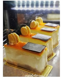 Three beautifully decorated slices of cake are displayed in a well-lit setting. Each slice has a smooth, creamy texture and is topped with a layer of orange glaze, a single macaron, and a small piece of dark chocolate. The cakes rest on golden bases, emphasizing a luxurious presentation.