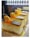 Three beautifully decorated slices of cake are displayed in a well-lit setting. Each slice has a smooth, creamy texture and is topped with a layer of orange glaze, a single macaron, and a small piece of dark chocolate. The cakes rest on golden bases, emphasizing a luxurious presentation.