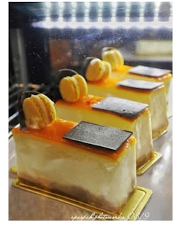 Three beautifully decorated slices of cake are displayed in a well-lit setting. Each slice has a smooth, creamy texture and is topped with a layer of orange glaze, a single macaron, and a small piece of dark chocolate. The cakes rest on golden bases, emphasizing a luxurious presentation.