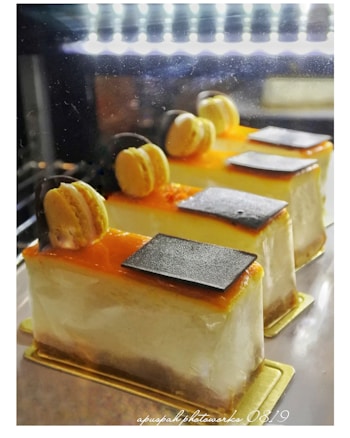 Three beautifully decorated slices of cake are displayed in a well-lit setting. Each slice has a smooth, creamy texture and is topped with a layer of orange glaze, a single macaron, and a small piece of dark chocolate. The cakes rest on golden bases, emphasizing a luxurious presentation.