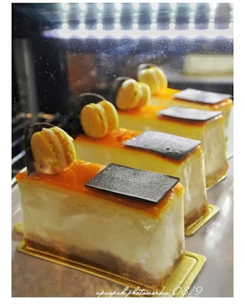 Three beautifully decorated slices of cake are displayed in a well-lit setting. Each slice has a smooth, creamy texture and is topped with a layer of orange glaze, a single macaron, and a small piece of dark chocolate. The cakes rest on golden bases, emphasizing a luxurious presentation.