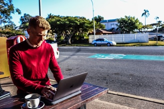 A man wearing a red sweater is working on a laptop at an outdoor table. A cup of coffee sits nearby. The background shows a street with parked cars and a few trees, suggesting a suburban setting.