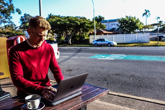 A man wearing a red sweater is working on a laptop at an outdoor table. A cup of coffee sits nearby. The background shows a street with parked cars and a few trees, suggesting a suburban setting.