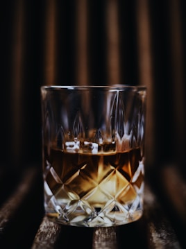 A close-up of a glass filled with an amber-colored liquid, likely whiskey, on a dark wooden surface. The glass has an intricate cut pattern that reflects light, creating a sophisticated and elegant atmosphere.