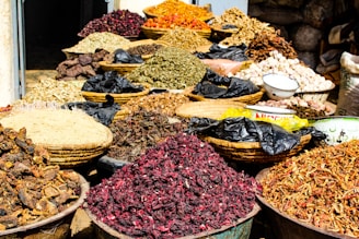 A variety of dried herbs and spices displayed in large woven baskets and bowls, creating a vibrant and colorful scene. The spices range in color, with shades of red, brown, green, and orange, and are piled high, showcasing an abundance of natural ingredients. The setting suggests a market or bazaar atmosphere.