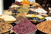 A variety of dried herbs and spices displayed in large woven baskets and bowls, creating a vibrant and colorful scene. The spices range in color, with shades of red, brown, green, and orange, and are piled high, showcasing an abundance of natural ingredients. The setting suggests a market or bazaar atmosphere.