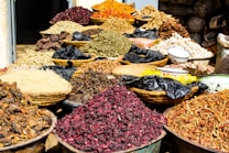 A variety of dried herbs and spices displayed in large woven baskets and bowls, creating a vibrant and colorful scene. The spices range in color, with shades of red, brown, green, and orange, and are piled high, showcasing an abundance of natural ingredients. The setting suggests a market or bazaar atmosphere.