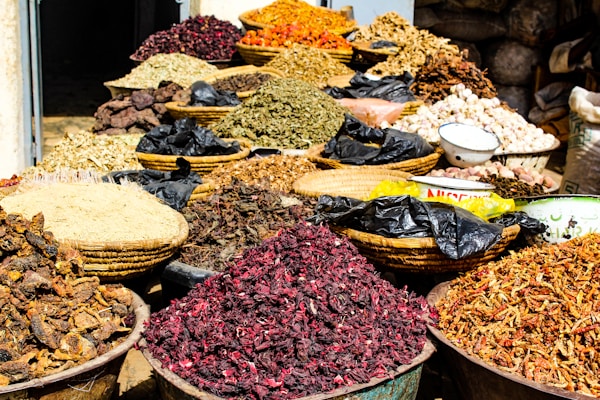 A variety of dried herbs and spices displayed in large woven baskets and bowls, creating a vibrant and colorful scene. The spices range in color, with shades of red, brown, green, and orange, and are piled high, showcasing an abundance of natural ingredients. The setting suggests a market or bazaar atmosphere.
