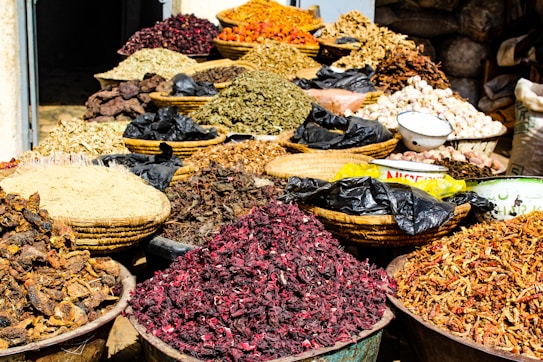 A variety of dried herbs and spices displayed in large woven baskets and bowls, creating a vibrant and colorful scene. The spices range in color, with shades of red, brown, green, and orange, and are piled high, showcasing an abundance of natural ingredients. The setting suggests a market or bazaar atmosphere.
