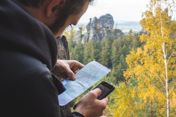 A person is examining a map and using a smartphone in an outdoor setting surrounded by trees. In the background, there are rock formations and a forested landscape. The trees appear to be in autumn colors with hues of yellow and green.