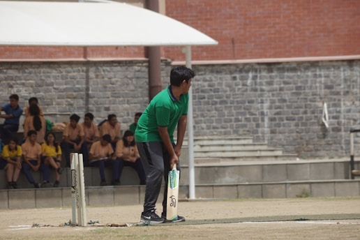 A person wearing a green shirt is preparing to bat on a cricket field. In the background, several people are seated on concrete steps, some wearing yellow and others wearing beige shirts. The setting appears to be an outdoor area with a stone wall and an overhang providing shade.
