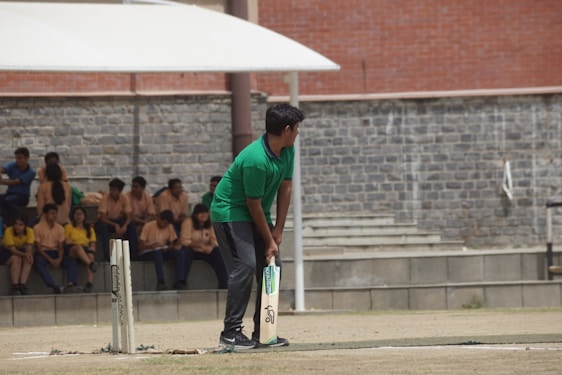 A person wearing a green shirt is preparing to bat on a cricket field. In the background, several people are seated on concrete steps, some wearing yellow and others wearing beige shirts. The setting appears to be an outdoor area with a stone wall and an overhang providing shade.