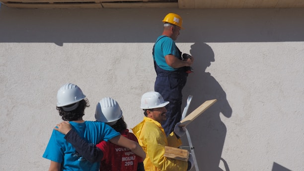A group of four people wearing hard hats stands near a wall. One person on a ladder, wearing a yellow helmet and holding a power drill, is working on the wall. Another member of the group is carrying wooden planks. The others are dressed in colorful t-shirts and are facing the wall, possibly discussing or observing the work being done.