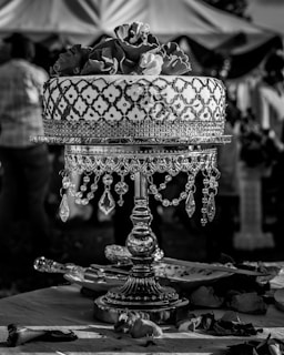 An intricately designed cake sits atop an ornate stand adorned with hanging crystals. The cake is decorated with a pattern and topped with realistic flower designs. In the background, silhouettes of people and draped fabric suggest an event setting. Rose petals are scattered on the table around the cake stand.