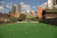 An empty urban soccer field with artificial green turf is surrounded by a high net. In the background, there are tall residential and commercial buildings, set against a clear blue sky with scattered white clouds. The scene suggests a recreational area within a city environment.