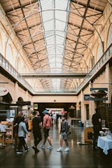 A bustling indoor public space with high ceilings and large windows, featuring a variety of shops along the sides. People are casually walking and interacting, indicating a lively atmosphere. The architecture is industrial, with exposed beams and a spacious layout.