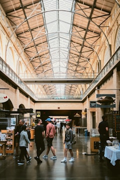 A bustling indoor public space with high ceilings and large windows, featuring a variety of shops along the sides. People are casually walking and interacting, indicating a lively atmosphere. The architecture is industrial, with exposed beams and a spacious layout.