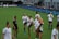 A group of female soccer players in matching uniforms is stretching on a soccer field. They are practicing high kicks as part of their warm-up routine. The grass is bright green, and there are small orange training cones scattered around. A coach or trainer in black attire is observing their practice.
