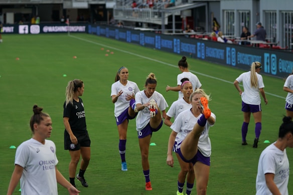 A group of female soccer players in matching uniforms is stretching on a soccer field. They are practicing high kicks as part of their warm-up routine. The grass is bright green, and there are small orange training cones scattered around. A coach or trainer in black attire is observing their practice.