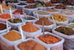 An assortment of bags filled with various colorful spices are displayed, each containing a metal spoon. The spices range from bright orange, red, and yellow powders to whole spices like cinnamon sticks. The bags are placed in close proximity, creating a vibrant and aromatic market scene.