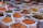 An assortment of bags filled with various colorful spices are displayed, each containing a metal spoon. The spices range from bright orange, red, and yellow powders to whole spices like cinnamon sticks. The bags are placed in close proximity, creating a vibrant and aromatic market scene.