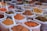 An assortment of bags filled with various colorful spices are displayed, each containing a metal spoon. The spices range from bright orange, red, and yellow powders to whole spices like cinnamon sticks. The bags are placed in close proximity, creating a vibrant and aromatic market scene.