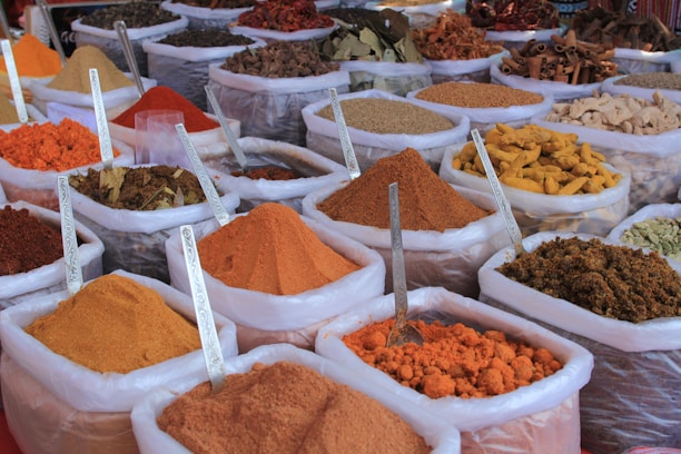 An assortment of bags filled with various colorful spices are displayed, each containing a metal spoon. The spices range from bright orange, red, and yellow powders to whole spices like cinnamon sticks. The bags are placed in close proximity, creating a vibrant and aromatic market scene.