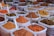 An assortment of bags filled with various colorful spices are displayed, each containing a metal spoon. The spices range from bright orange, red, and yellow powders to whole spices like cinnamon sticks. The bags are placed in close proximity, creating a vibrant and aromatic market scene.