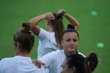 A group of women in white sports attire are on a green field. One woman, with her hair in a bun, looks directly at the camera, while another is seen from the back, holding a shirt. Another is in the background tying her hair.