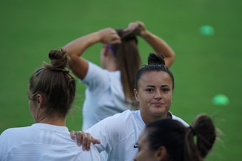 A group of women in white sports attire are on a green field. One woman, with her hair in a bun, looks directly at the camera, while another is seen from the back, holding a shirt. Another is in the background tying her hair.