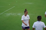 A woman wearing a white sports jersey with 'Orlando Health' written on it stands on a grassy field, smiling and engaging with another person who is partially visible. She has her hair tied up and is wearing shorts. The field is well-maintained and appears to be an athletic or sports setting.
