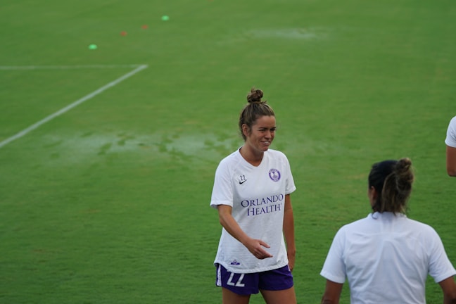 A woman wearing a white sports jersey with 'Orlando Health' written on it stands on a grassy field, smiling and engaging with another person who is partially visible. She has her hair tied up and is wearing shorts. The field is well-maintained and appears to be an athletic or sports setting.