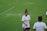 A woman wearing a white sports jersey with 'Orlando Health' written on it stands on a grassy field, smiling and engaging with another person who is partially visible. She has her hair tied up and is wearing shorts. The field is well-maintained and appears to be an athletic or sports setting.