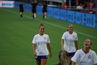 Young girls in colorful jerseys warming up on a sunny football field.