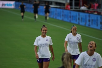 Young girls in colorful jerseys warming up on a sunny football field.