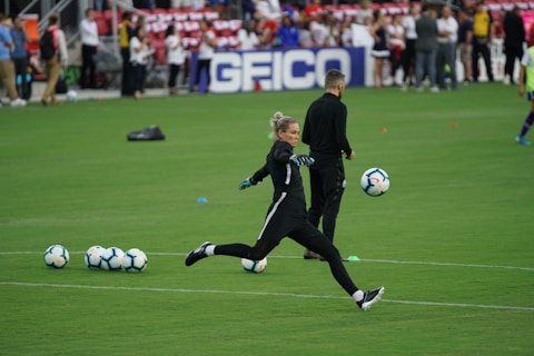 A soccer player in a black sports outfit is mid-action on a green field, focusing intently on kicking a soccer ball. Several soccer balls are scattered on the grass nearby, and a coach or teammate stands in the background watching. Spectators can be seen in the distant stands.