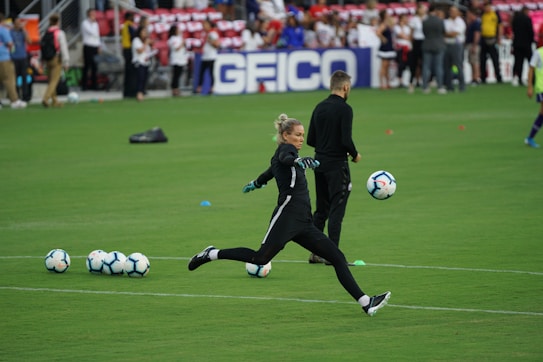 A soccer player in a black sports outfit is mid-action on a green field, focusing intently on kicking a soccer ball. Several soccer balls are scattered on the grass nearby, and a coach or teammate stands in the background watching. Spectators can be seen in the distant stands.