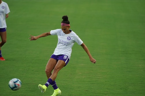 A soccer player in a white jersey and purple shorts is kicking a soccer ball on a green field. The player has a focused expression and is wearing a headband and neon green cleats. Another player is partially visible in the background.